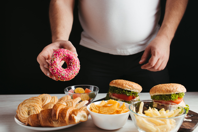 overweight man holding donut over assortment of junk food
