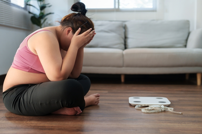 overweight woman sitting next to scale and tape measure with head in hands feeling frustrated